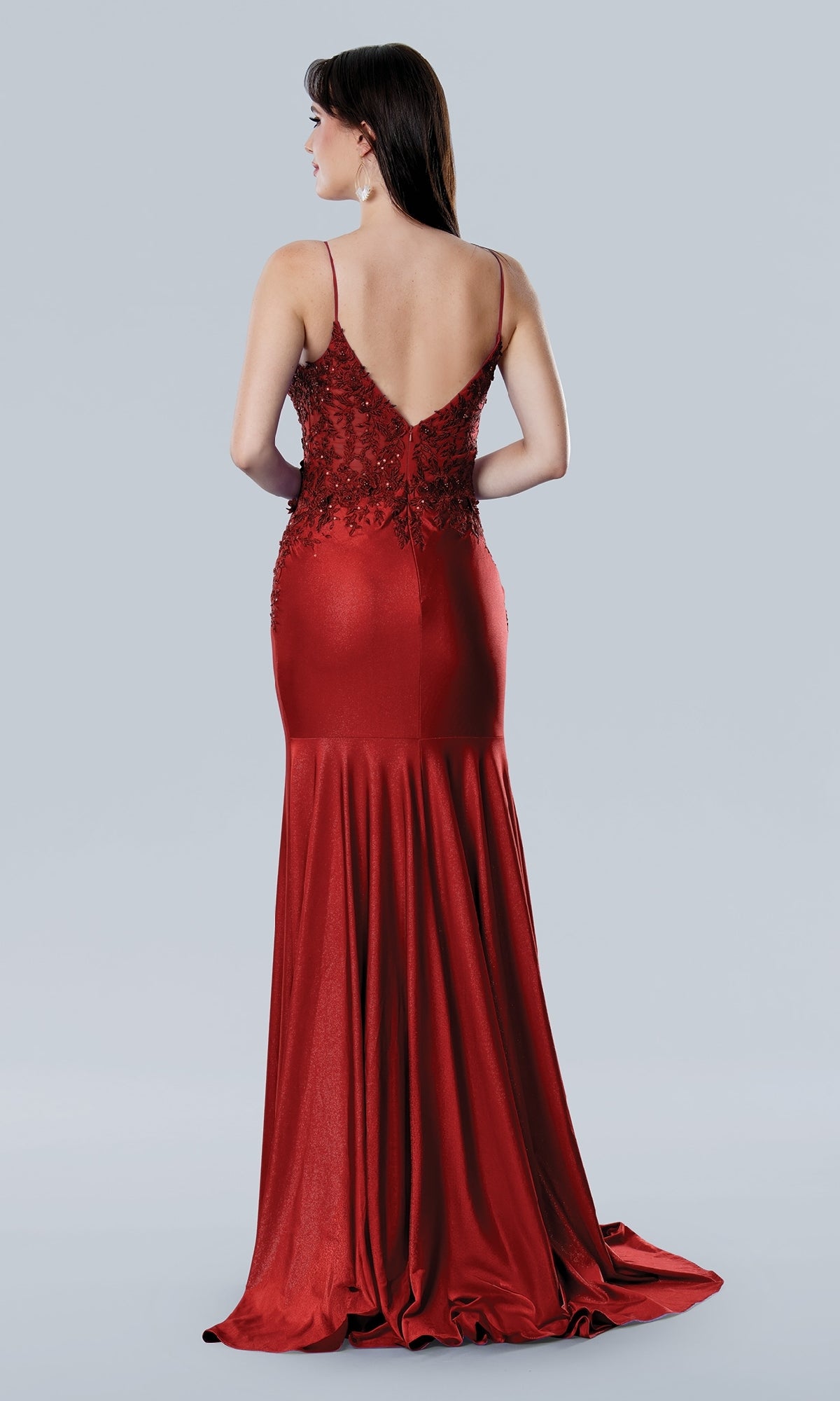 A woman wearing the Stella Couture 25186 long prom dress—a floor-length, sleeveless red sheath with a v-neckline and lace detailing—stands against a plain light background.
